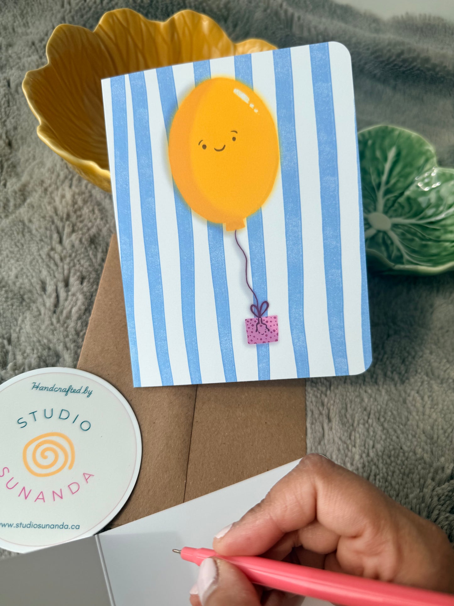 Hand writing a message on a cream card next to a cheerful balloon illustration
