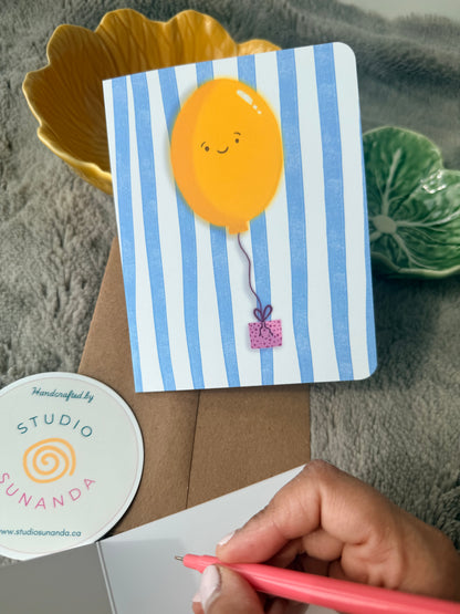 Hand writing a message on a cream card next to a cheerful balloon illustration